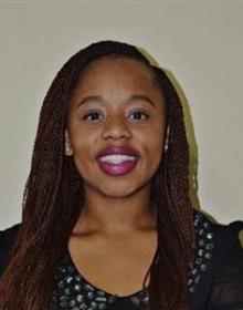 Headshot of Tuletu, a woman with long, dark red braids, wearing a black top with silver studs. She has bright pink li[ color and is smiling at the camera. The background is a plain beige wall, with a shadow cast behind her.