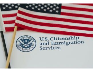 A graphic featuring two American flags, positioned behind a white background with text and a logo. The text reads U.S. Citizenship Immigration Services in blue font, accompanied by the Department of Homeland Security seal to the left.